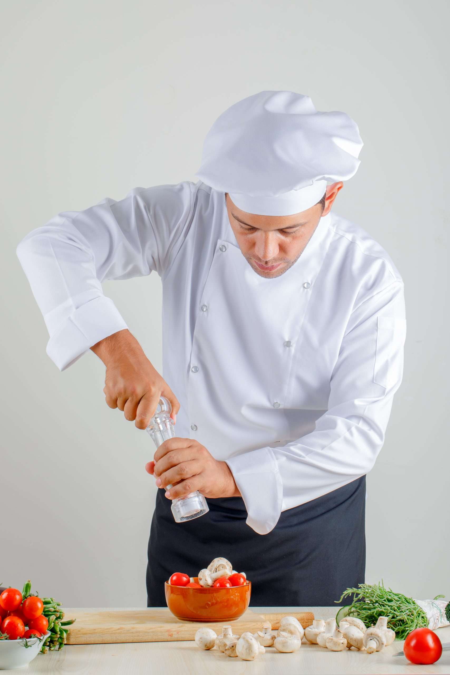 Male chef adding salt into food in uniform, hat and apron in kitchen , front view. Male chef adding salt into food in uniform, hat and apron in kitchen , front view.